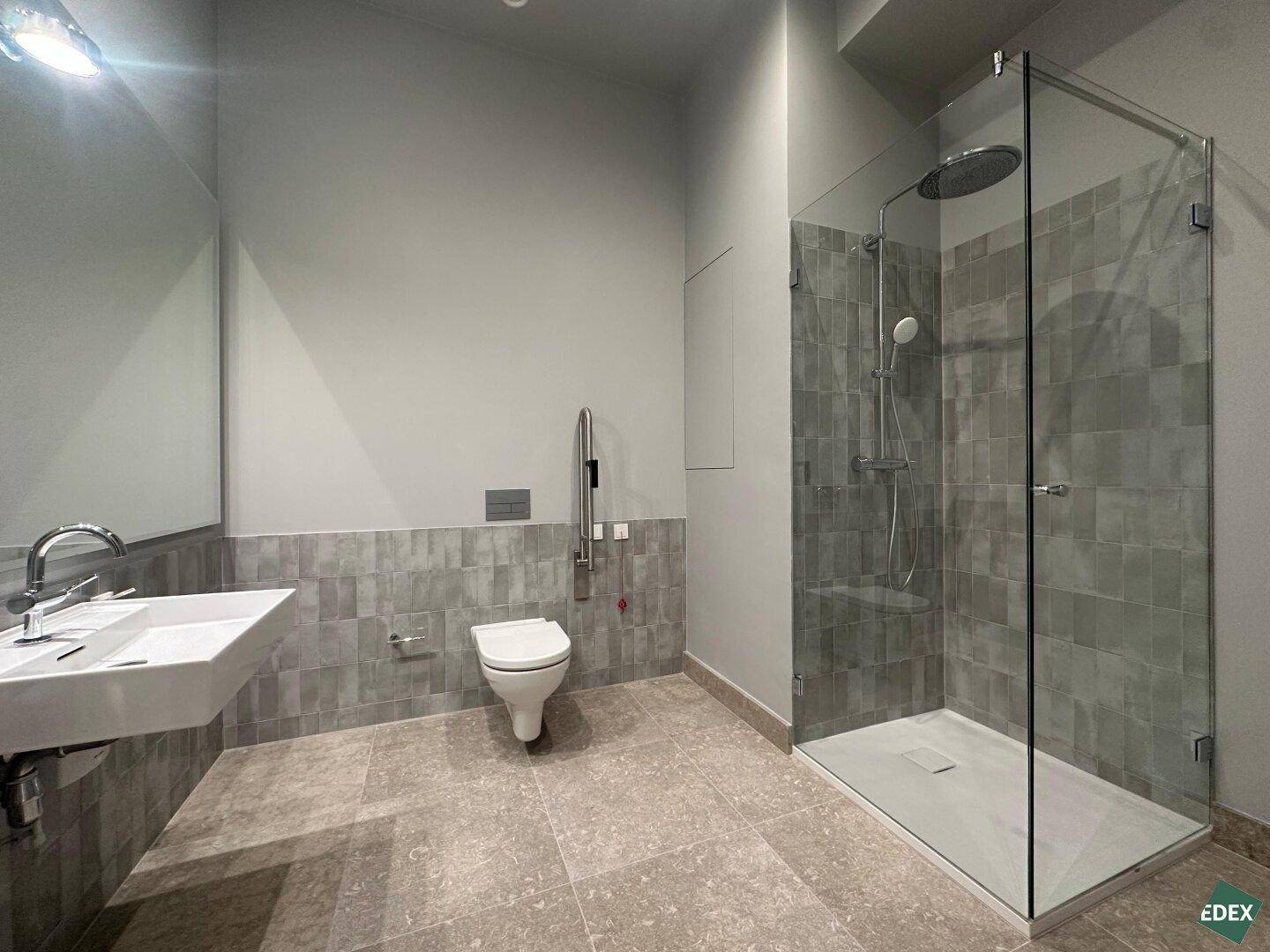 Modernes Badezimmer mit ebenerdiger Dusche, Wand-WC und großem Waschbecken, ausgestattet mit Haltegriffen.