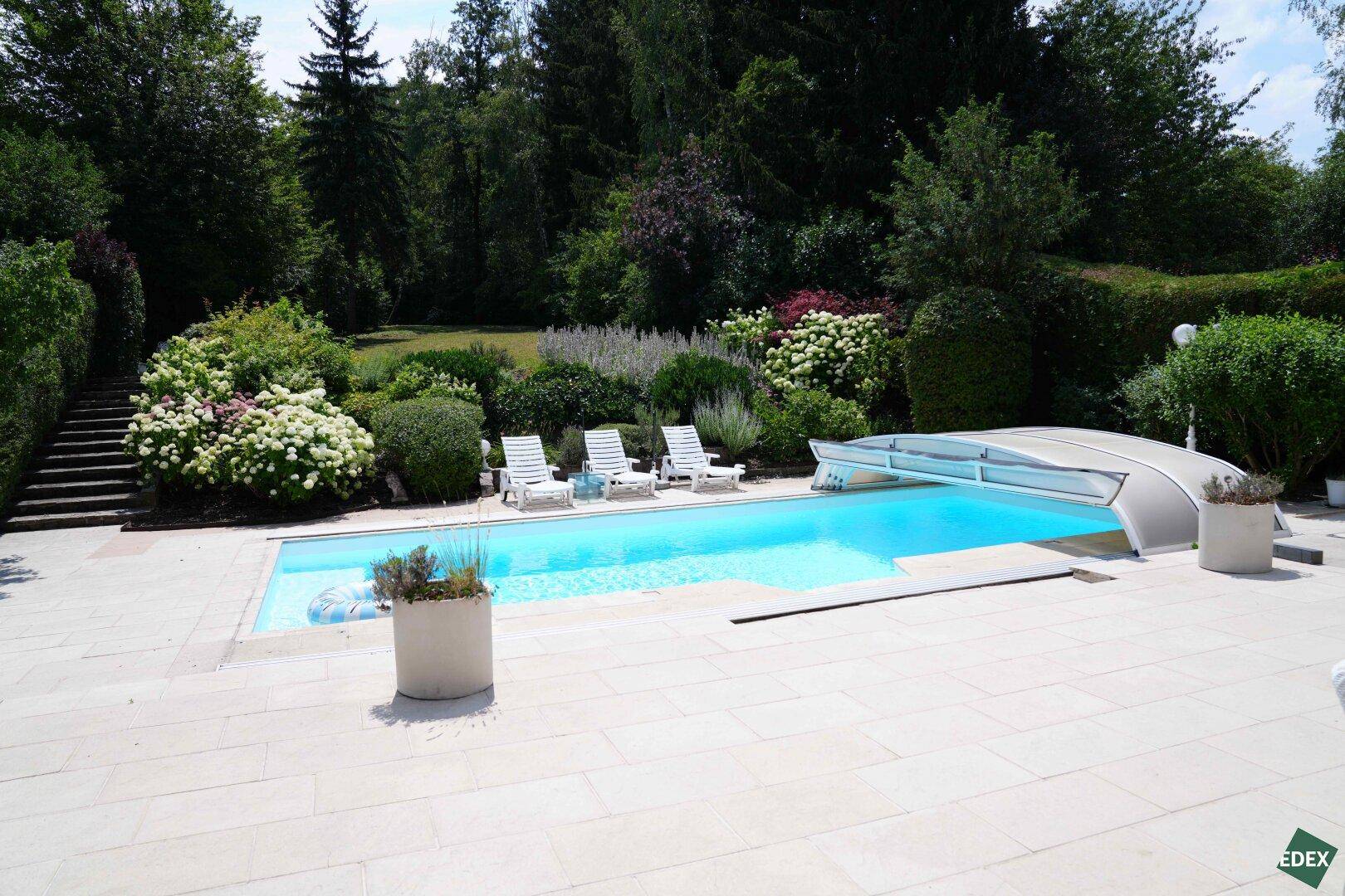 Großer Swimmingpool mit Abdeckung, umgeben von einer Terrasse und viel Grün.