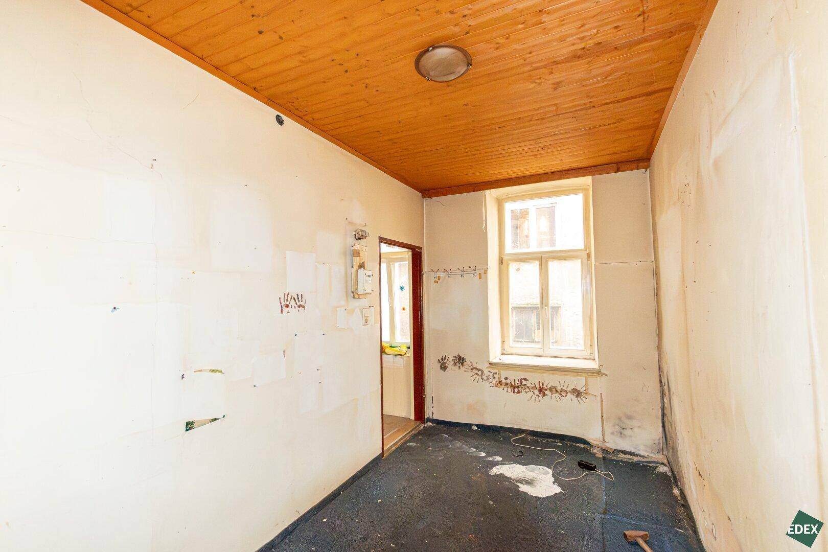 Kleiner, unrenovierter Raum mit Holzdecke, dunklem Boden und einem Fenster.