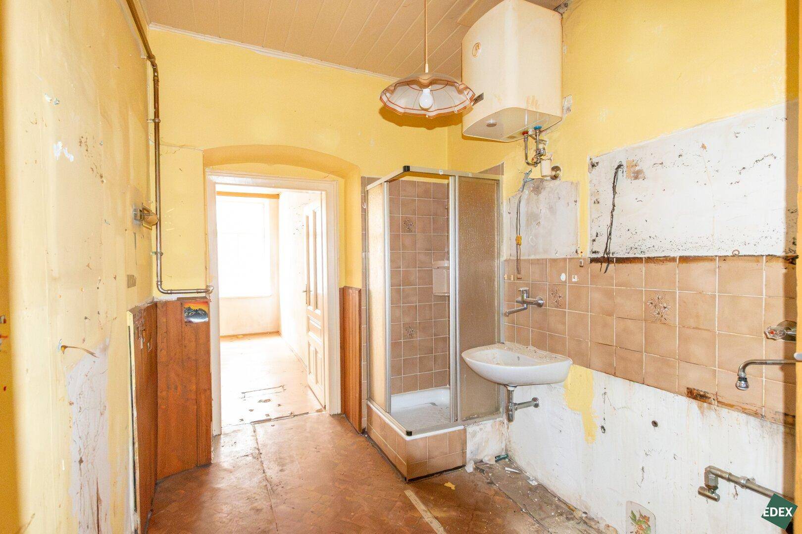 Sanierungsbedürftiges Badezimmer mit Duschkabine, Waschbecken, Fliesen und gelben Wänden.