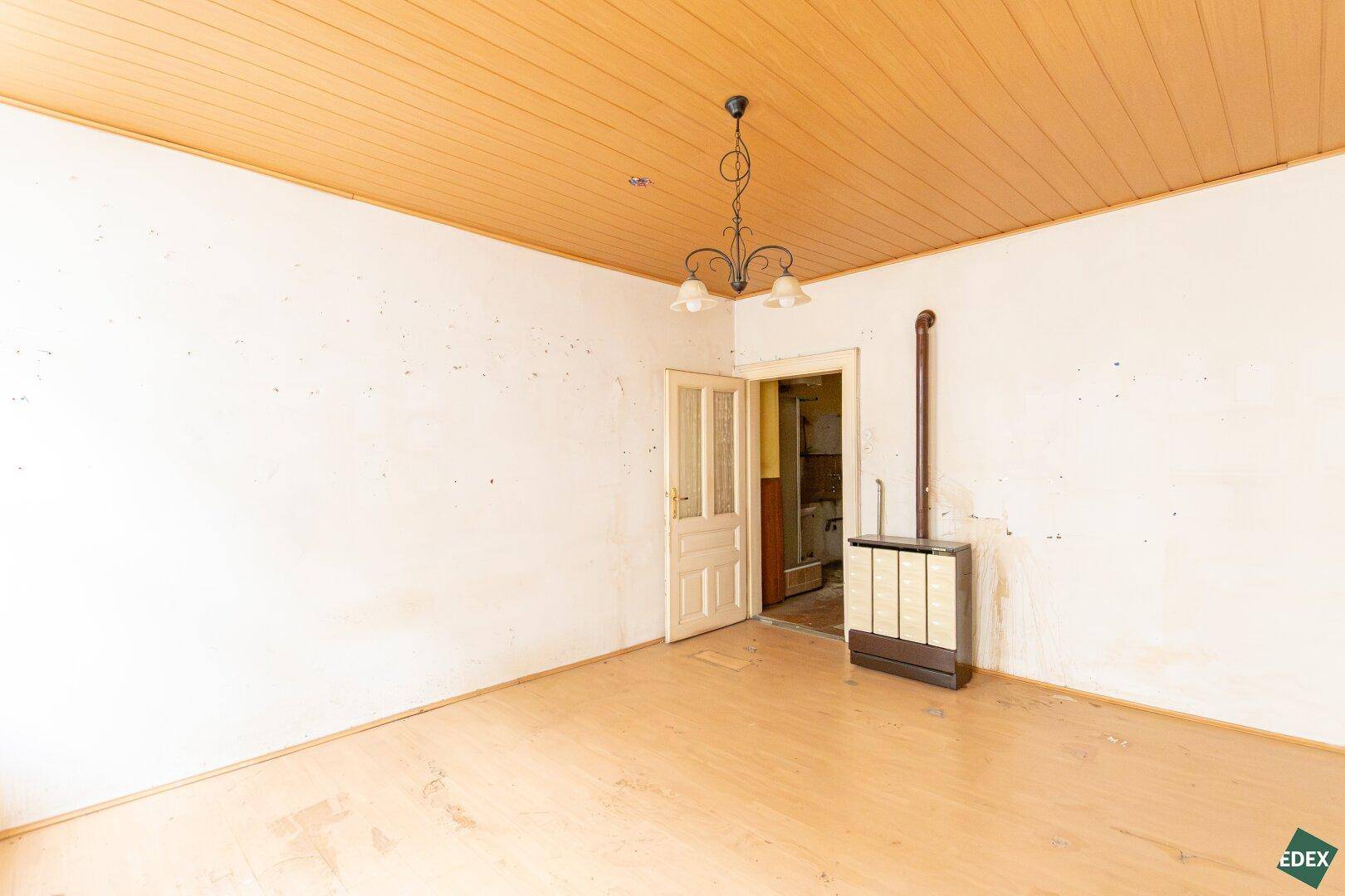Unrenovierter Raum mit Holzboden, Holzdecke und einem traditionellen Kachelofen.