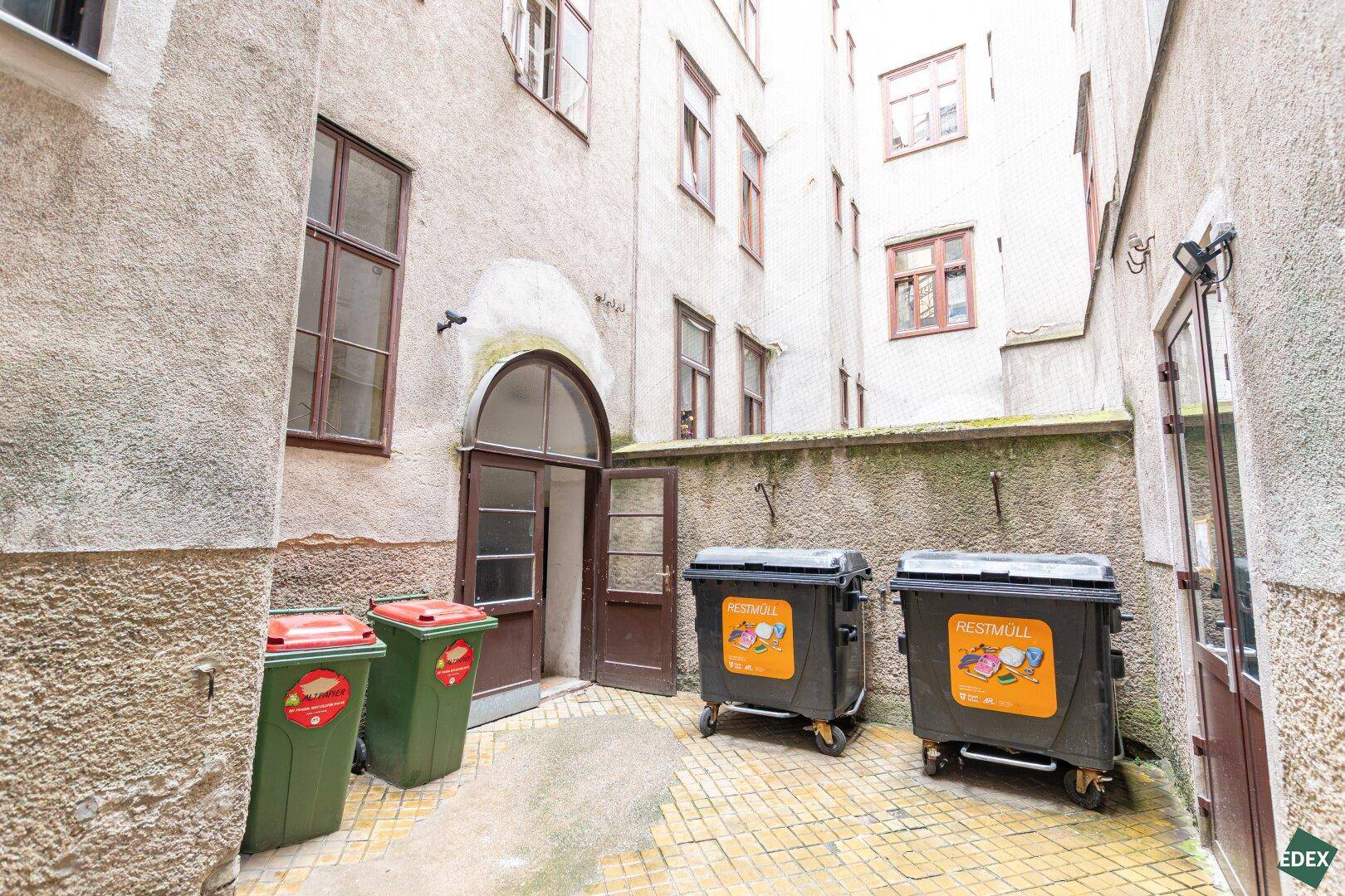 Innenhof eines Altbaus mit Mülltonnen und mehreren Fenstern, typisch für städtische Gebäude.
