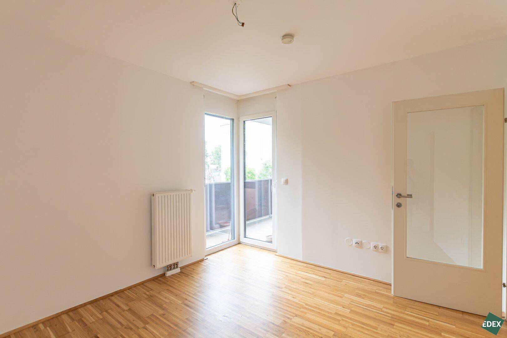Helles Zimmer mit Parkettboden und Eckfenstern, die Zugang zu einem Balkon bieten.