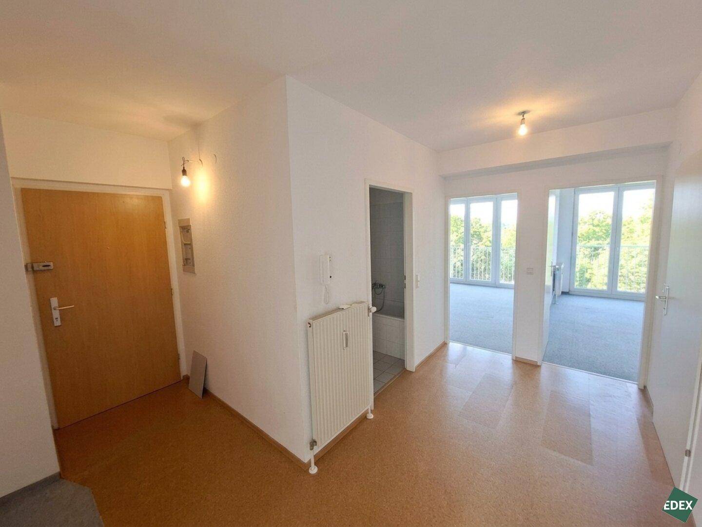 Heller Flur mit Korkboden, der zu einem Zimmer mit Teppichboden und großen Fenstern führt.
