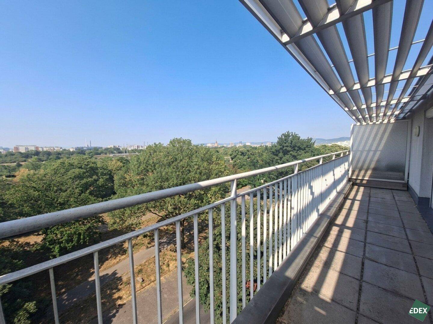 Langer Balkon mit Lamellendach und weitem Blick über grüne Bäume und die städtische Umgebung.