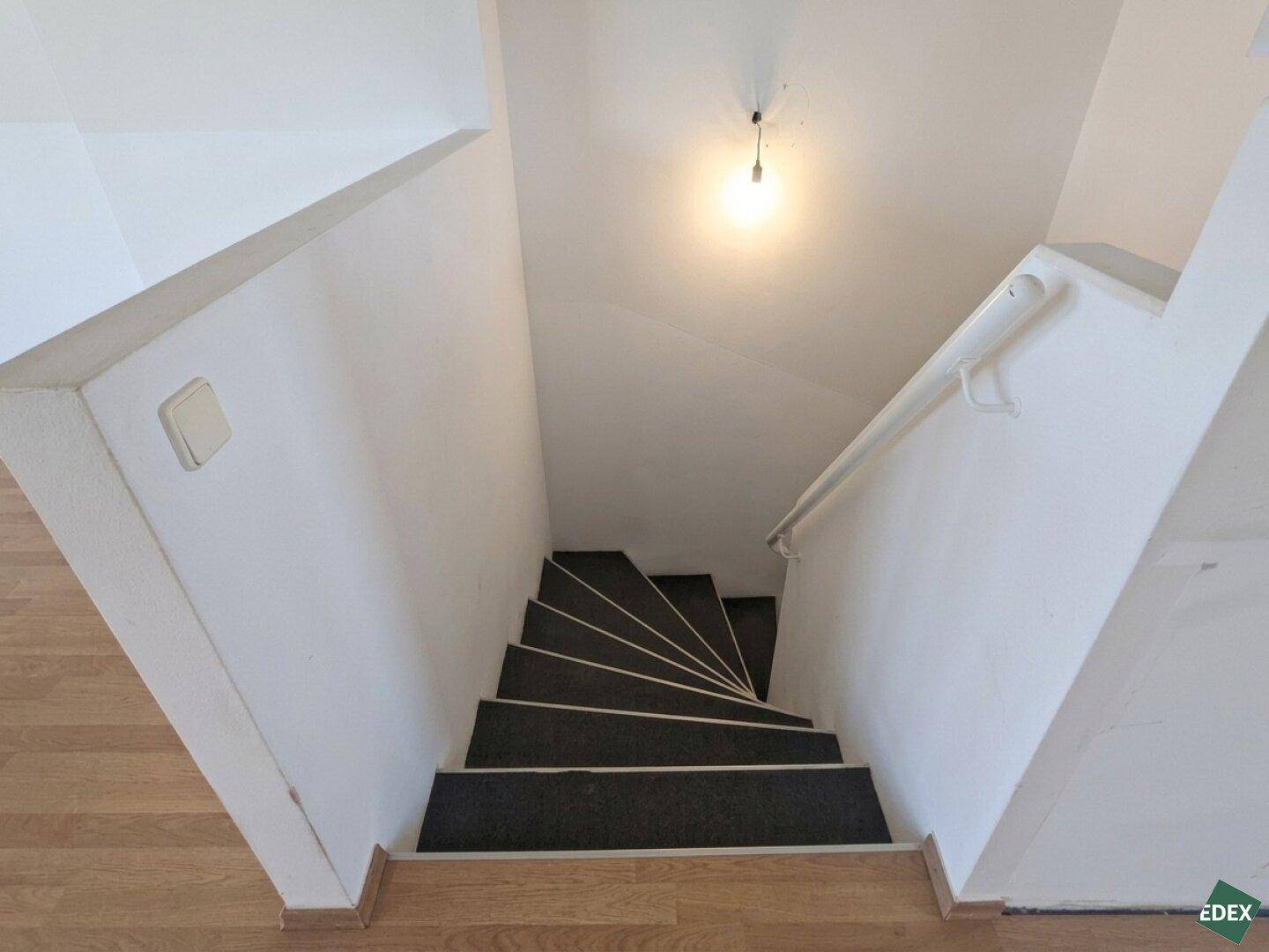 Innenliegende Treppe mit dunklen Stufen und weiß gestrichenen Wänden, die in eine andere Etage führt.