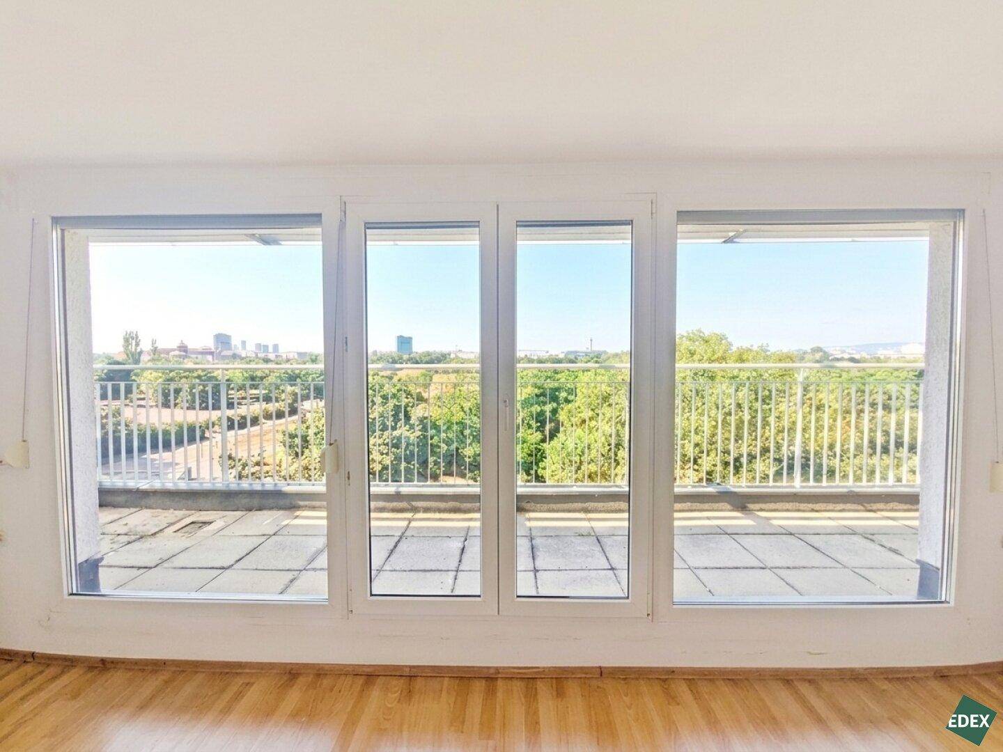 Große Fensterfront mit Zugang zu einer Terrasse und weitem Blick über Bäume und die Stadt.
