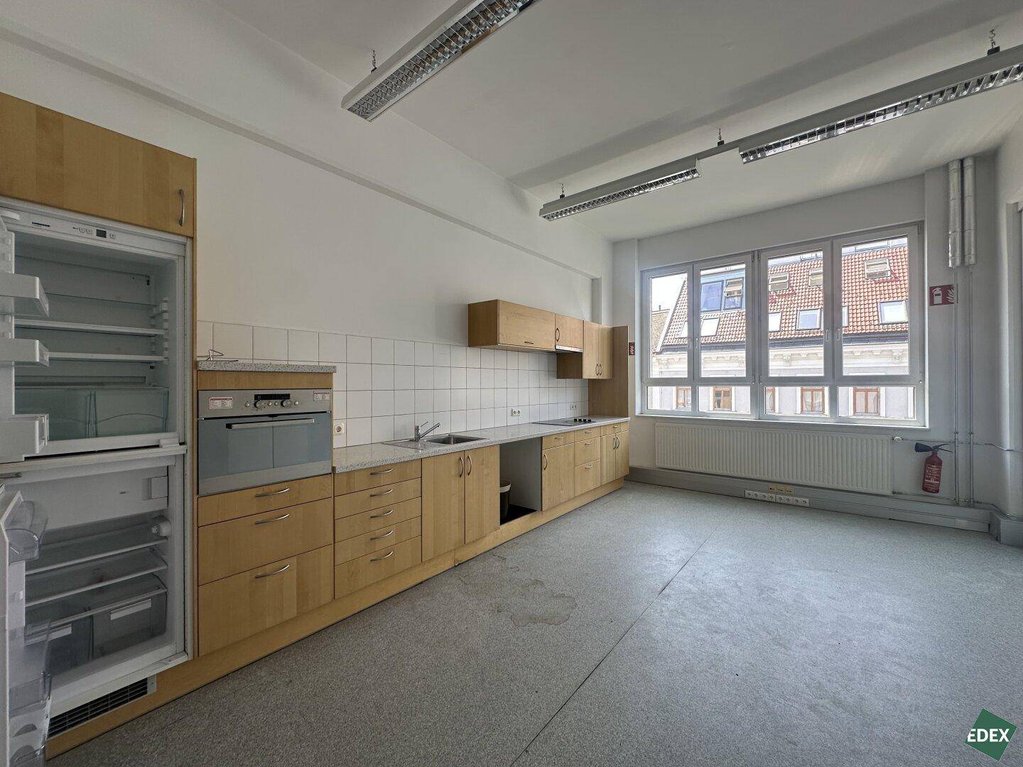 Funktionale Büroküche mit Holzschränken, Einbaugeräten und großen Fenstern, die viel Tageslicht hereinlassen.
