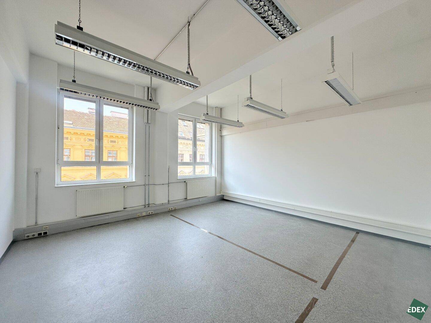 Leeres Büro mit zwei großen Fenstern, die viel Tageslicht hereinlassen, Betonboden und Heizkörpern unter den Fenstern.