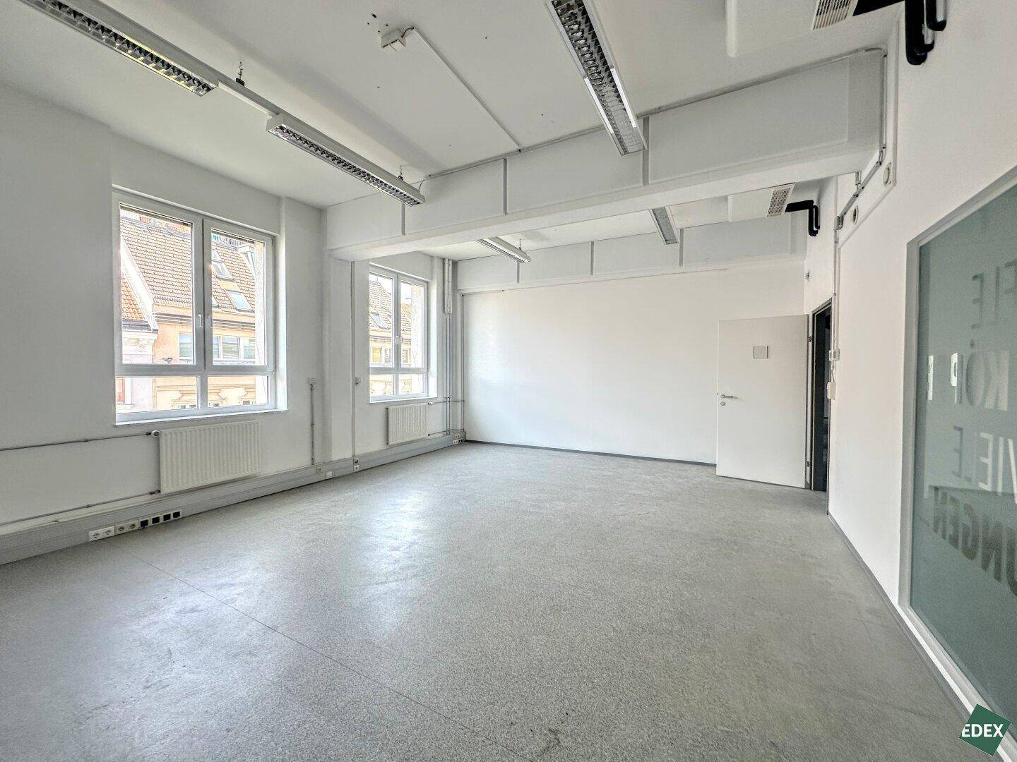 Geräumiges, leeres Büro mit großen Fenstern, Betonboden und einer Glaswand, die den Raum hell und offen wirken lässt.
