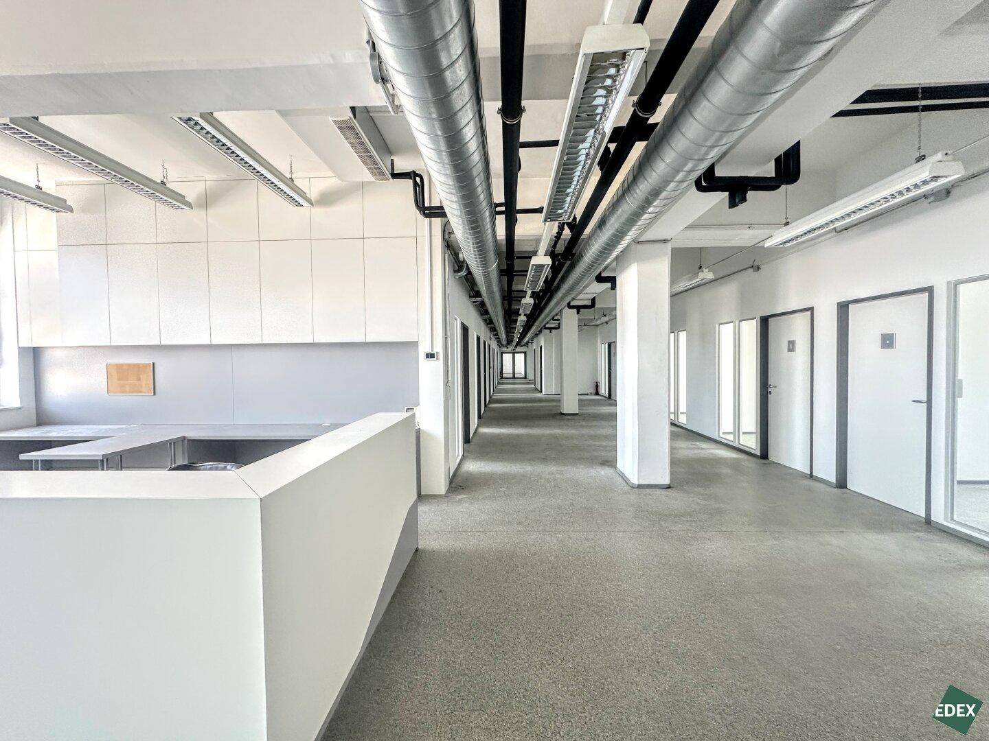 Langer Bürogang mit industriellem Charme, sichtbaren Lüftungsrohren und einem Empfangsbereich auf der linken Seite.