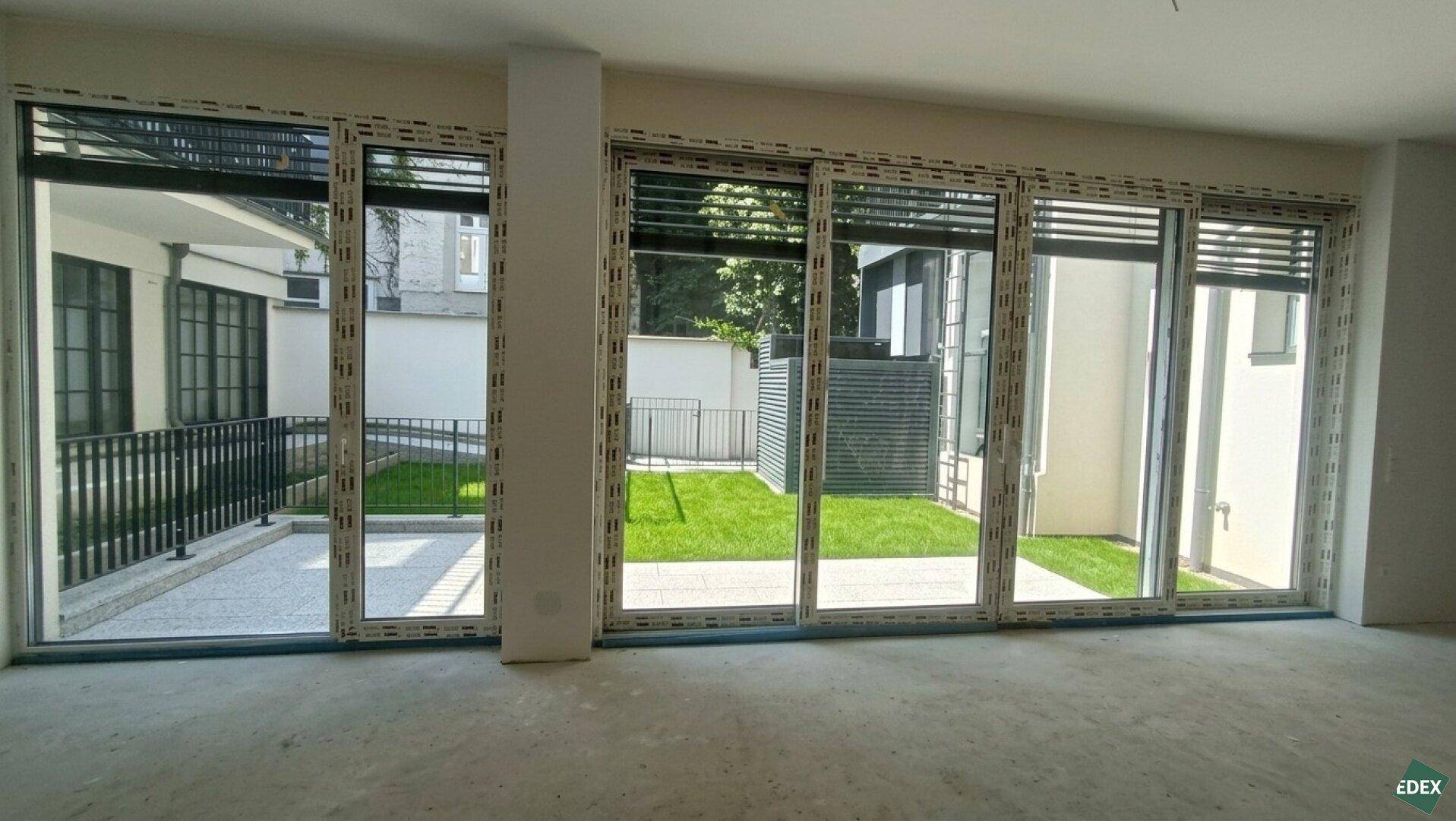 Große Fensterfront in einem unfertigen Raum mit Blick auf den begrünten Innenhof.