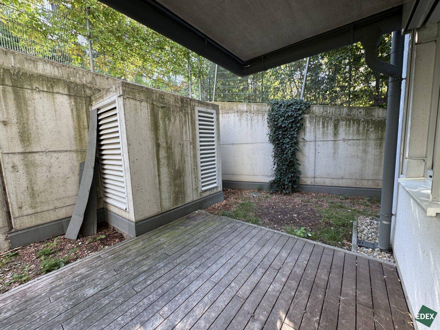 Holzterrasse im Freien neben einer Betonmauer mit Lüftungsschächten, bietet privaten Außenbereich.