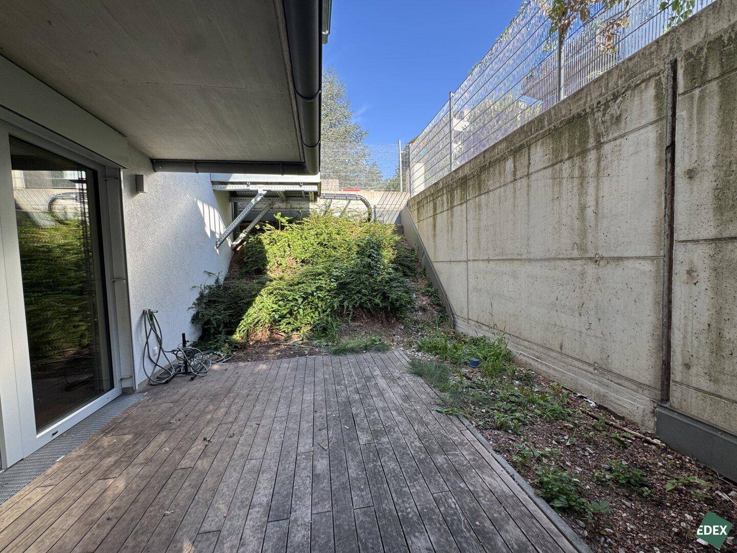 Holzterrasse im Freien neben einer Betonmauer mit üppigem Grün und Platz zum Entspannen.