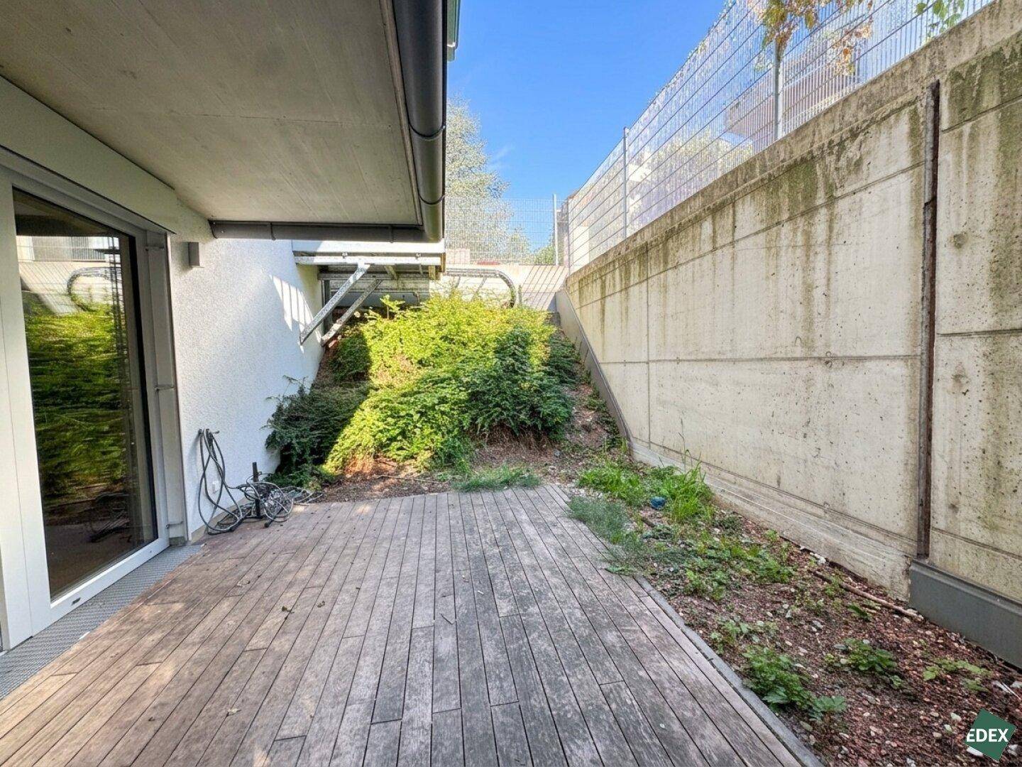 Holzterrasse im Freien neben einer Betonmauer mit üppigem Grün und Platz zum Entspannen.