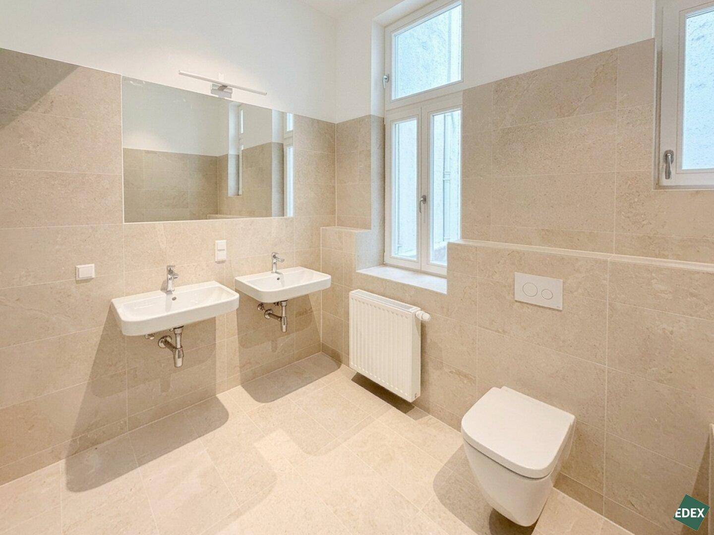 Modernes Badezimmer mit Doppelwaschbecken, Wand-WC und großen beigefarbenen Fliesen.