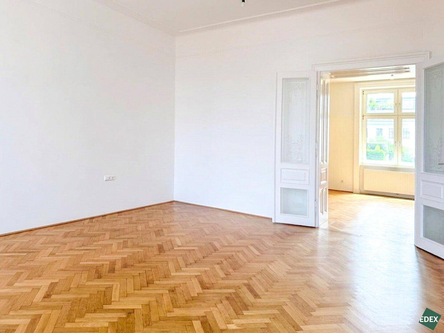 Geräumiger Wohnraum mit Fischgrätparkett und hohen Flügeltüren, die den Blick in ein helles Nebenzimmer freigeben.