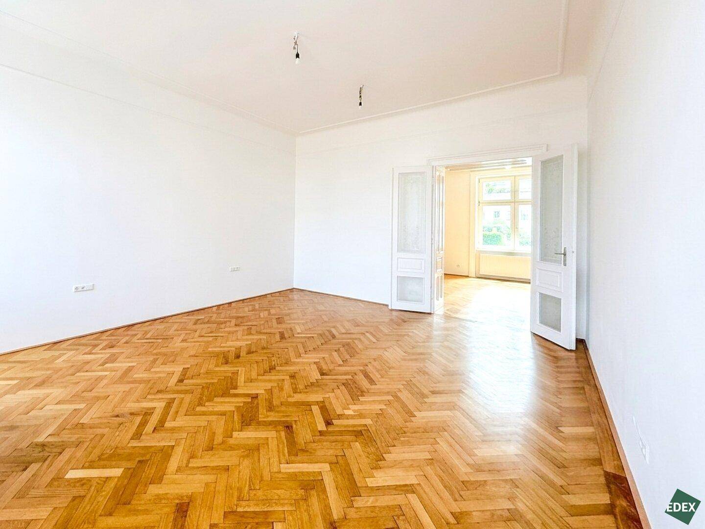 Heller Wohnbereich mit Fischgrätparkett und Flügeltüren, die den Zugang zu einem weiteren Zimmer ermöglichen.