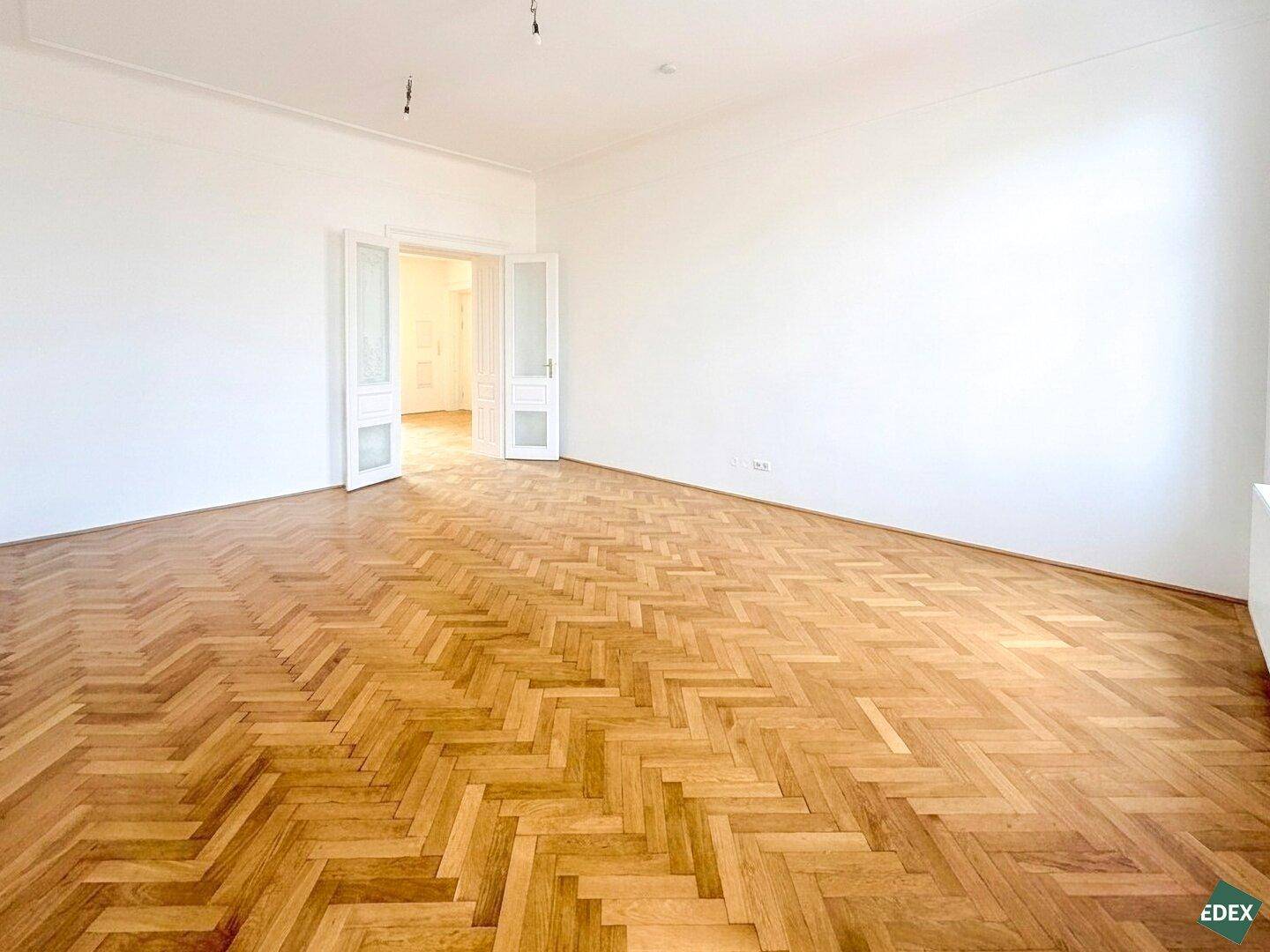 Eleganter Wohnraum mit Fischgrätparkett und hohen Flügeltüren, die zu einem angrenzenden Zimmer führen.