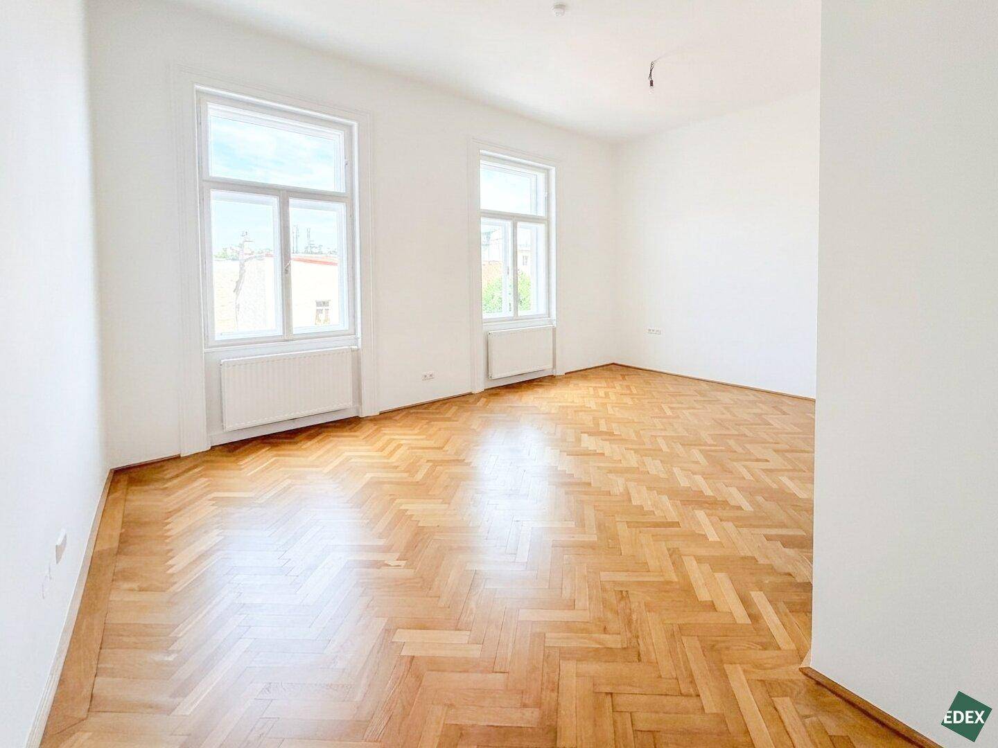 Heller und weitläufiger Raum mit klassischem Fischgrätparkett und großen Fenstern, die den Raum erhellen.