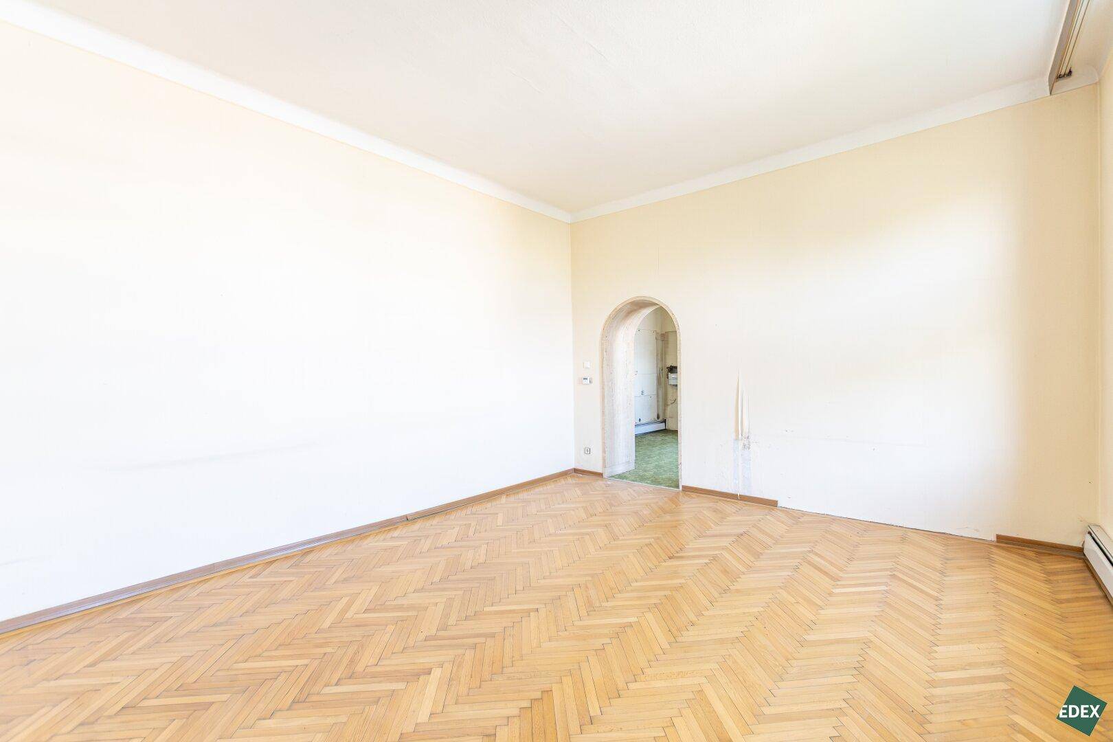 Geräumiges Zimmer mit Fischgrätparkett und einem Rundbogen, der in den nächsten Bereich führt.