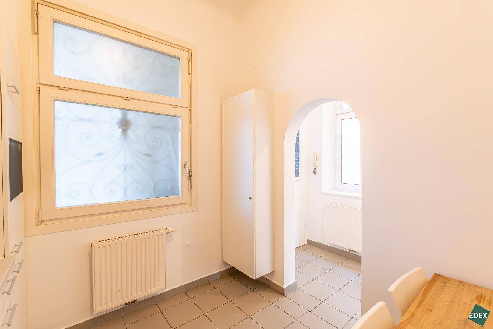 Heller Küchenbereich mit Fliesenboden, Fenster und einem Durchgang zu einem weiteren Raum.