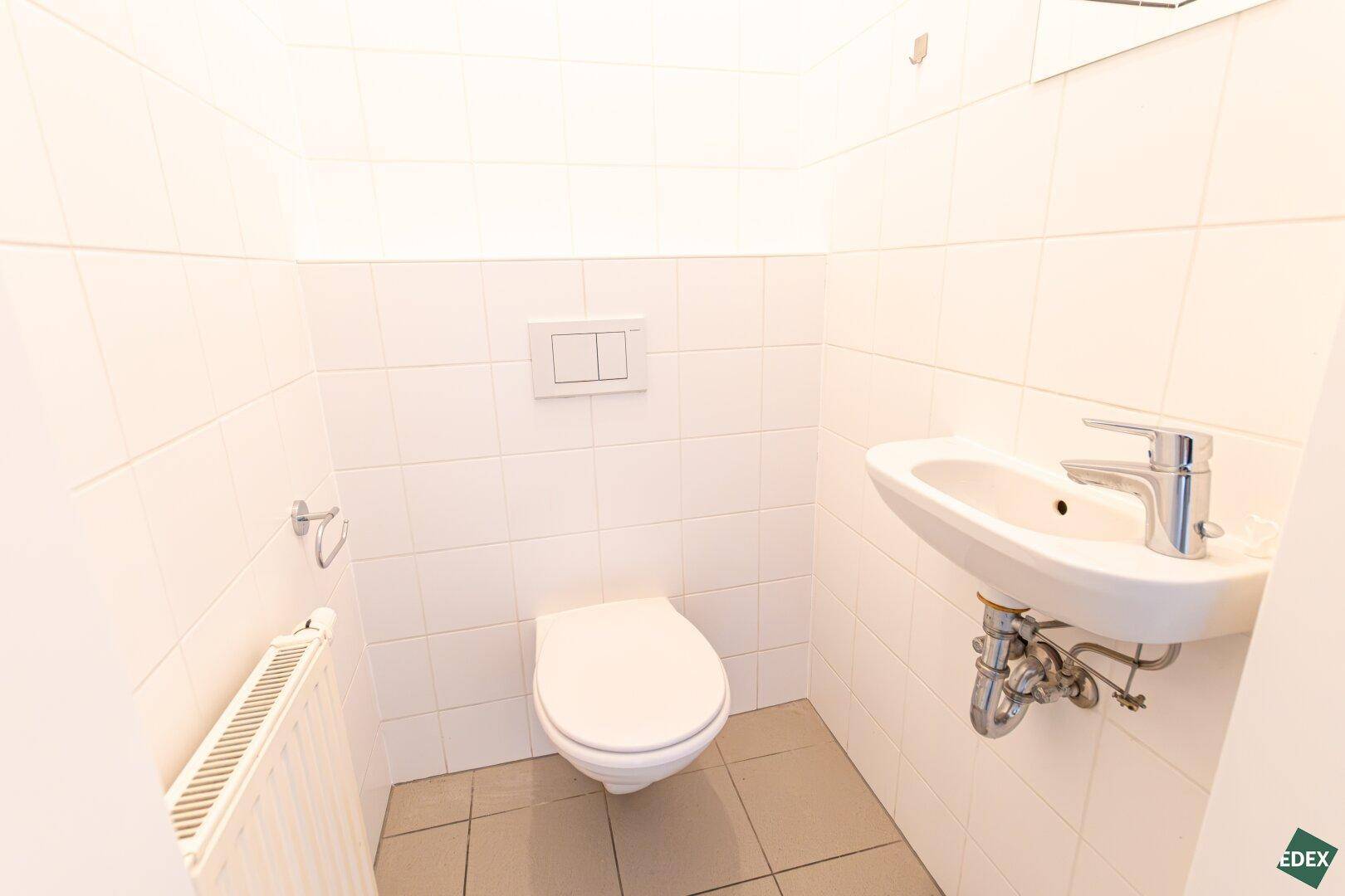 Separates WC mit Toilette, kleinem Waschbecken und weißen Fliesen, funktional und sauber.