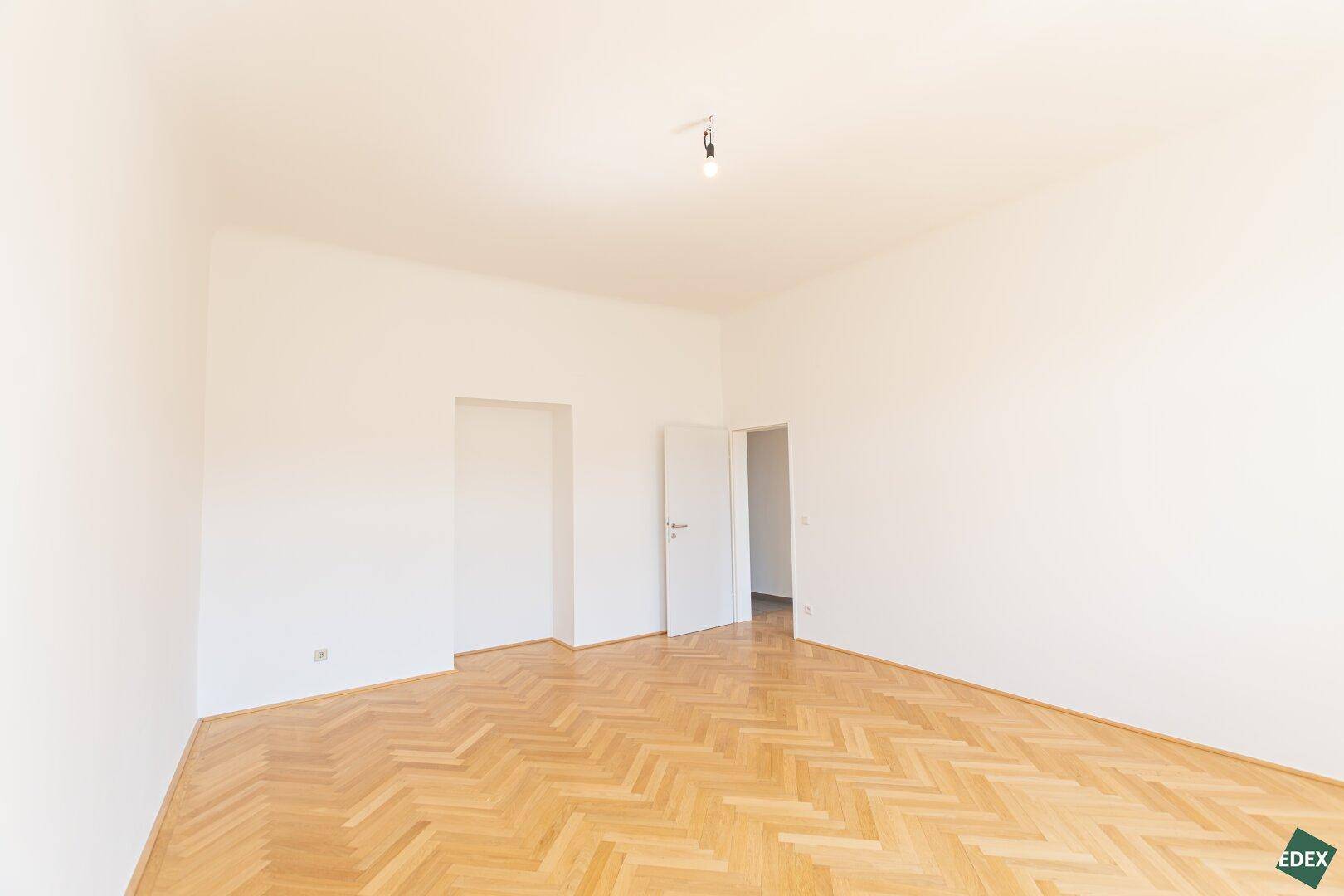Großes, leeres Zimmer mit Fischgrätparkett und zwei Türen, das viel Gestaltungspotenzial bietet.