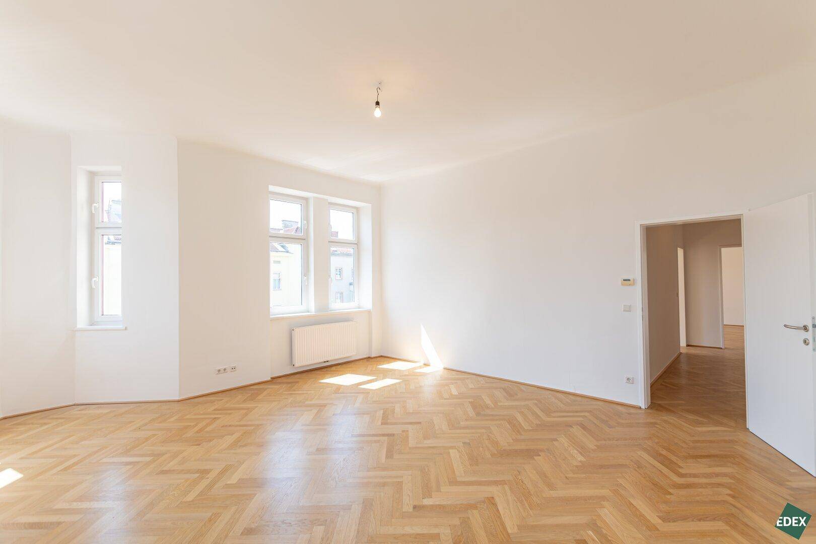 Großes, helles Zimmer mit Fischgrätparkett und mehreren Fenstern, das viel Tageslicht hereinlässt.