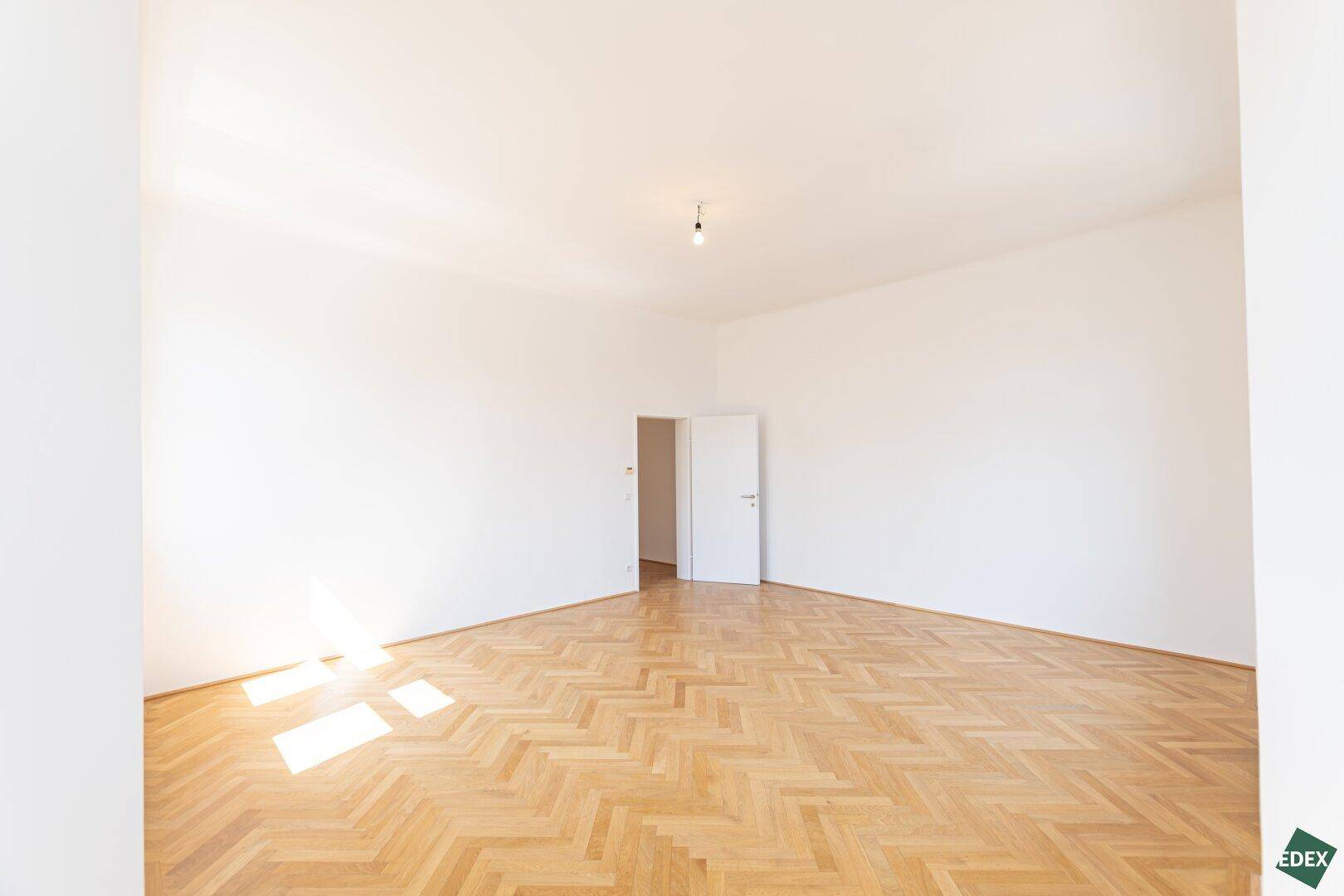 Geräumiges, leeres Zimmer mit Fischgrätparkett und weiß gestrichenen Wänden, bereit zur Gestaltung.
