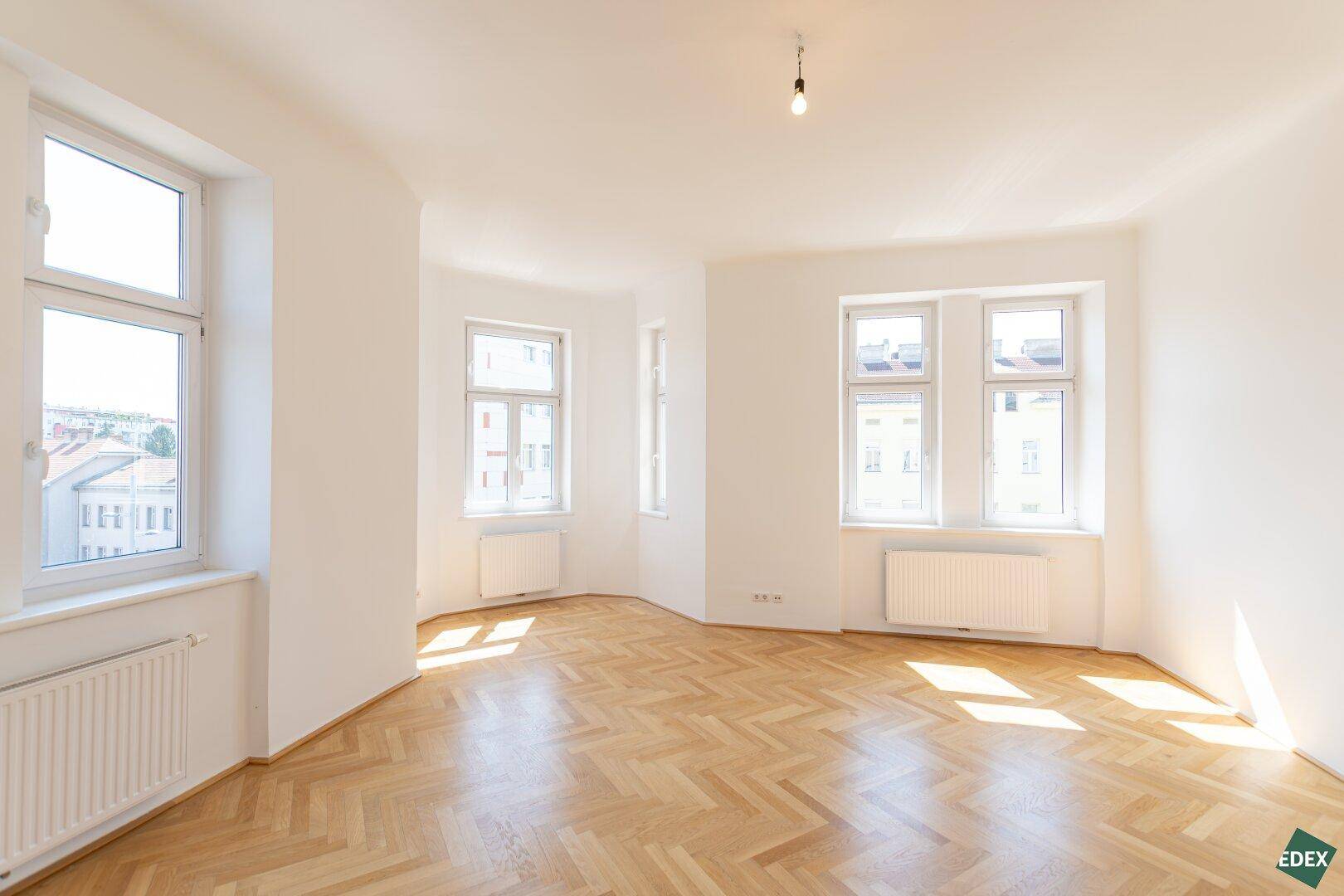 Helles, leeres Zimmer mit Fischgrätparkett und mehreren Fenstern, das viel Tageslicht hereinlässt.