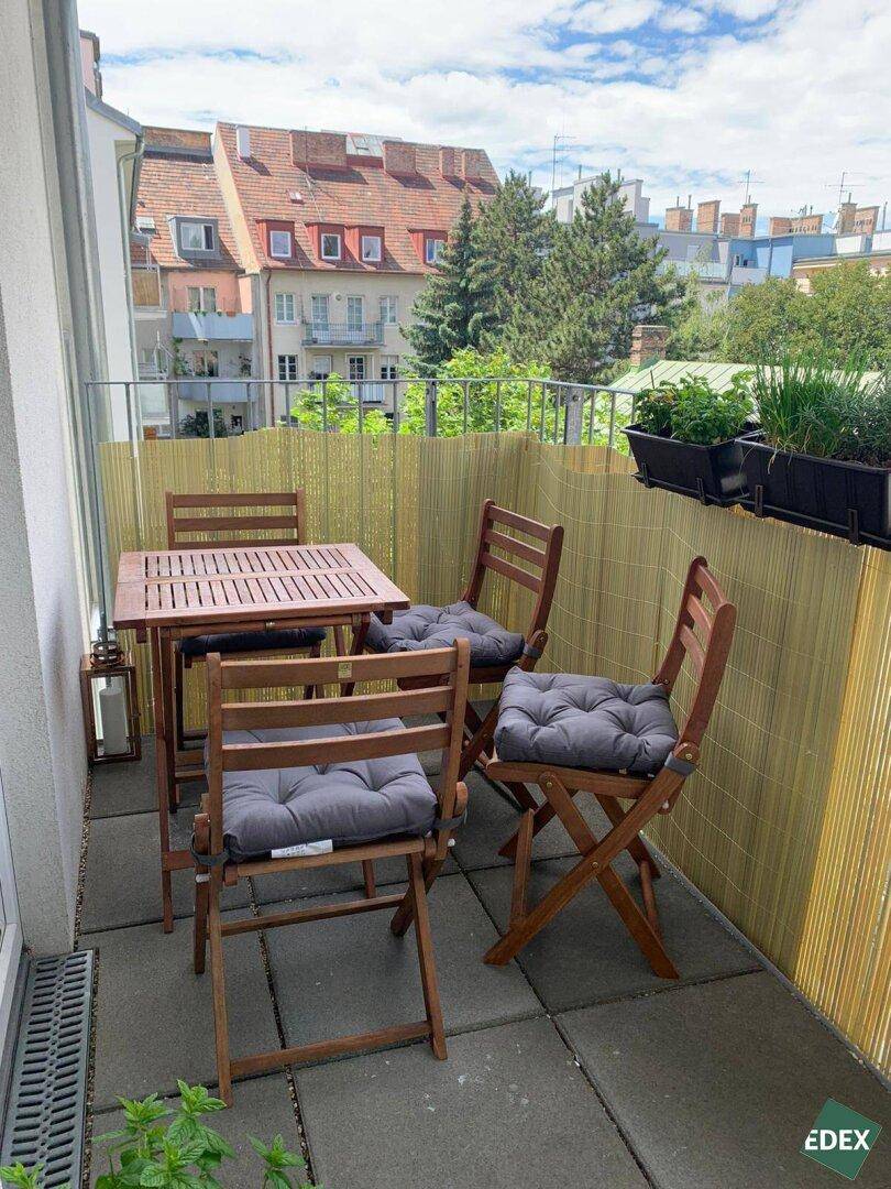 Gemütlicher Balkon mit Holztisch und Stühlen, umgeben von Pflanzen und mit Blick auf die Nachbarschaft.