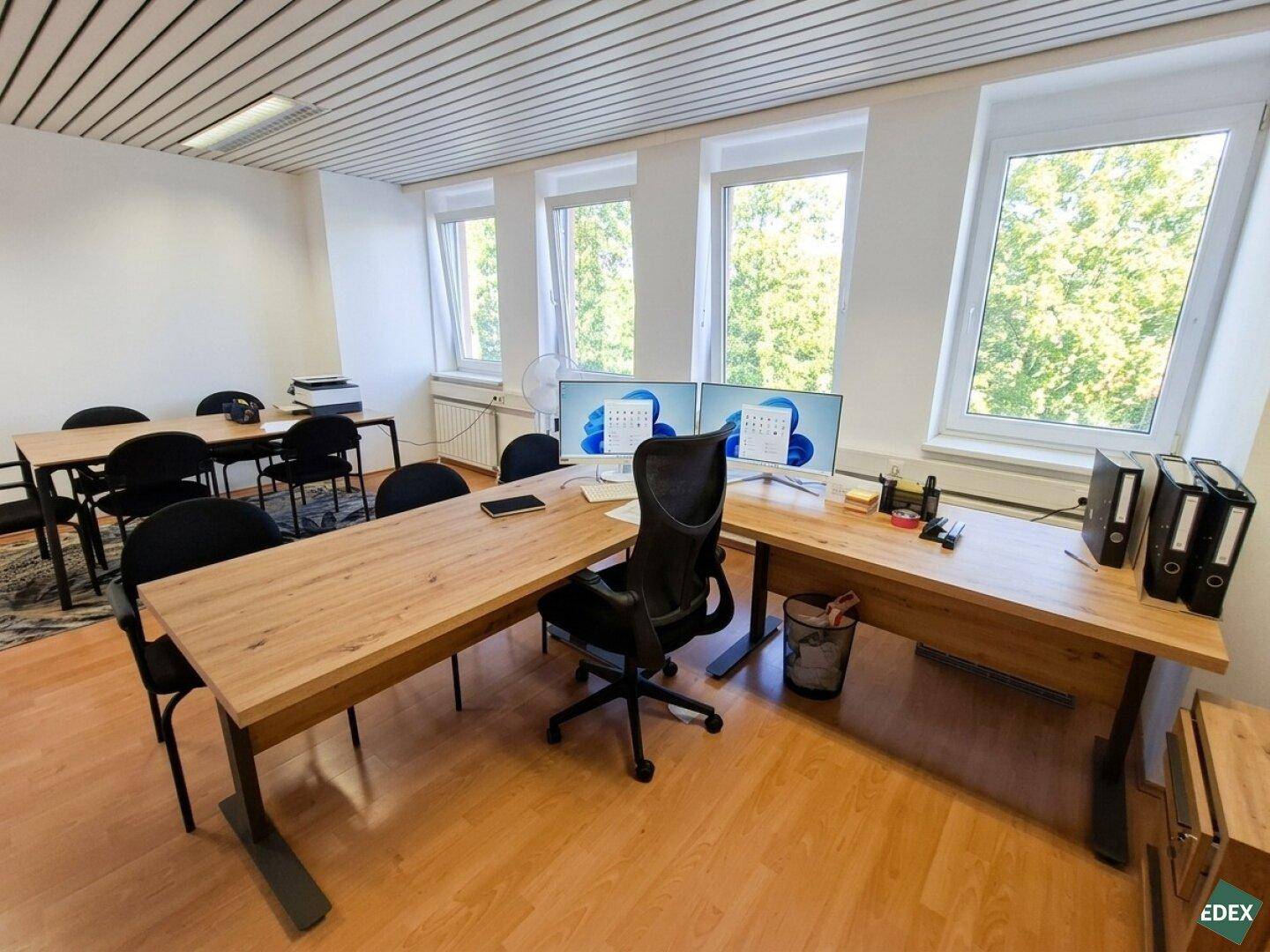 Geräumiges Büro mit zwei Monitoren auf einem Schreibtisch und Blick auf grüne Bäume.
