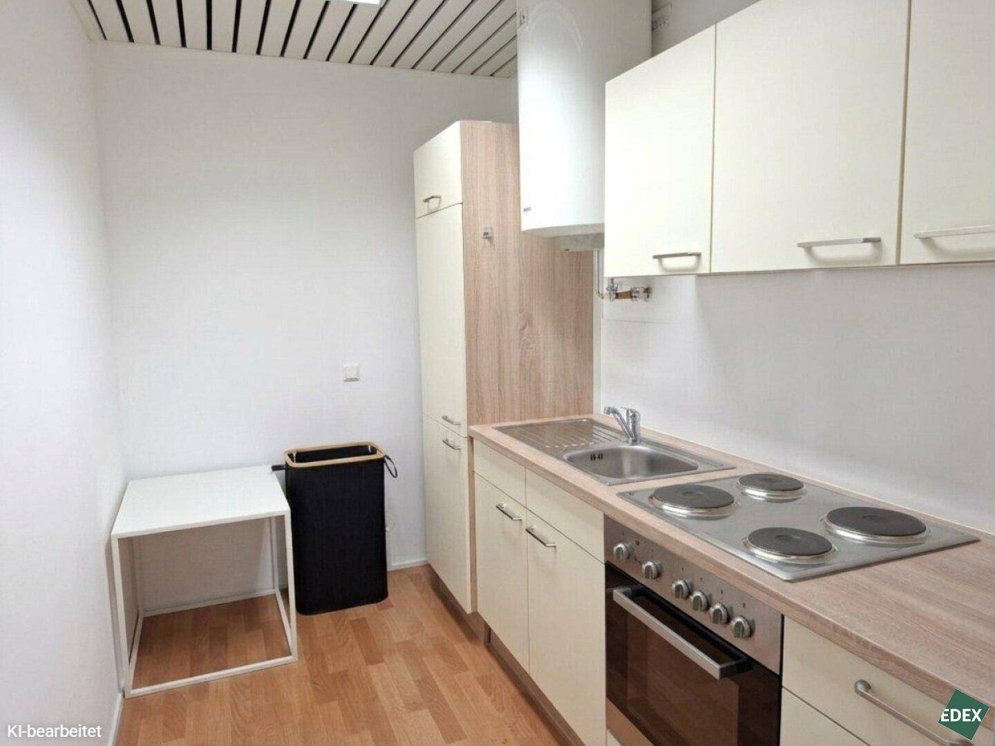 Kleine Küchenzeile mit Spüle, Herd, Backofen und hellen Schränken in einem funktionalen Raum.