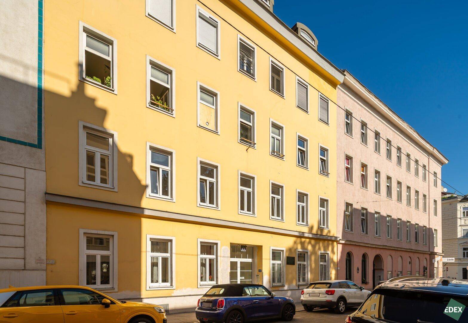 Gelbe Gebäudefassade an einer sonnigen Straße mit parkenden Autos und klarem blauem Himmel.