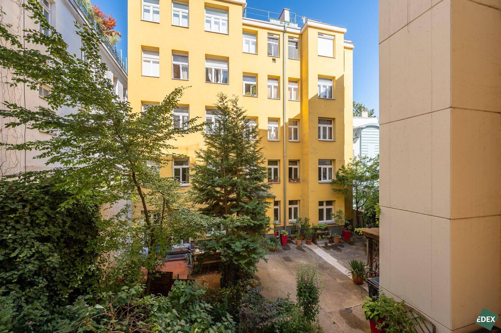 Blick auf den begrünten Innenhof mit hohen Bäumen und einer markanten gelben Gebäudefassade.