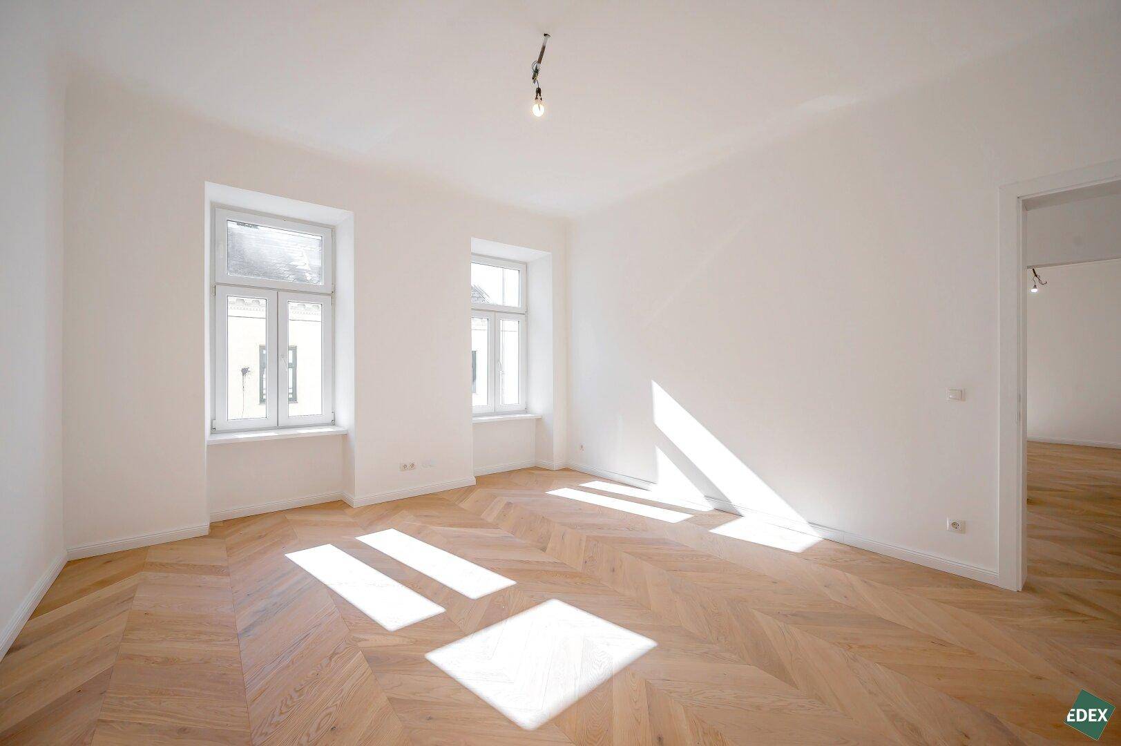 Lichtdurchflutetes Zimmer mit elegantem Fischgrätparkett und zwei hohen Fenstern.