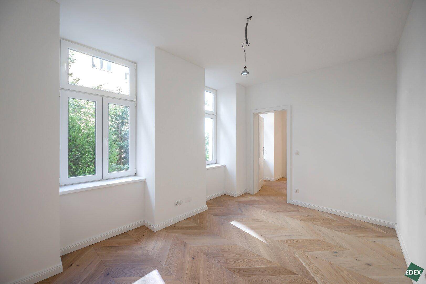 Helles Zimmer mit Fischgrätparkett und Blick ins Grüne durch die Fenster.