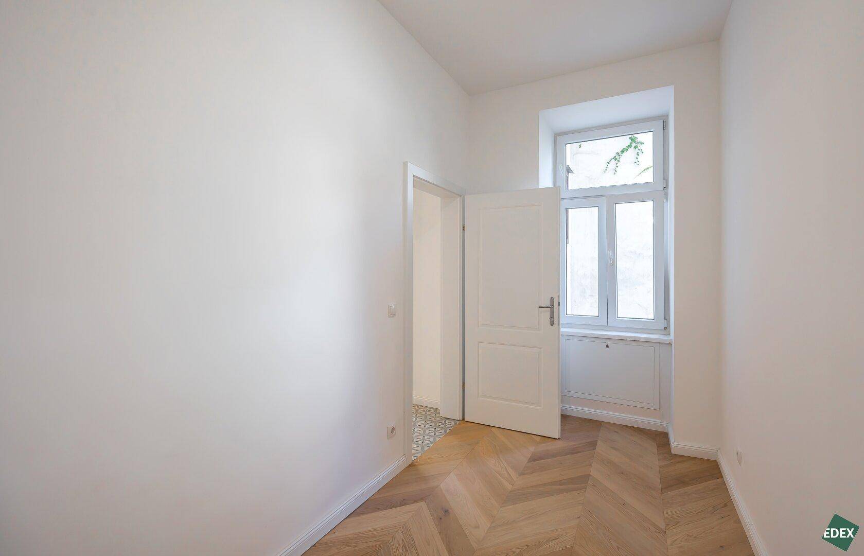 Helles Zimmer mit Fischgrätparkett und einem Fenster, das viel Tageslicht hereinlässt.