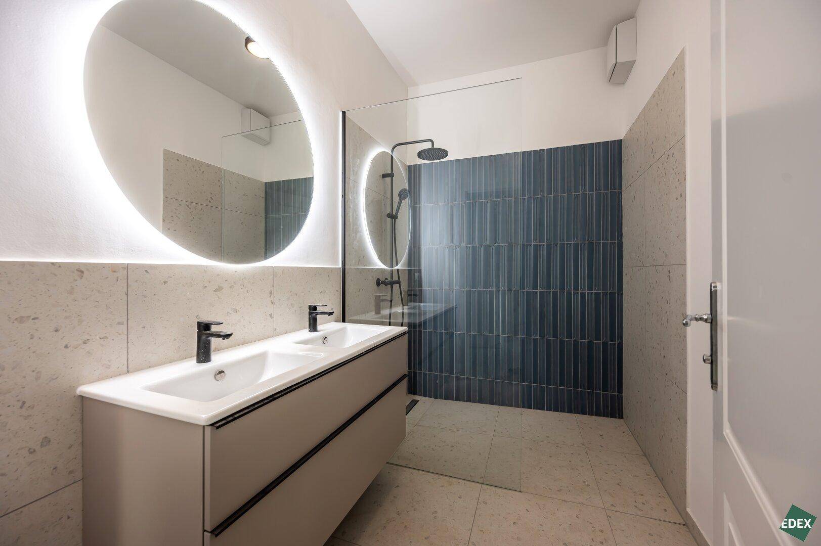 Modernes Badezimmer mit Doppelwaschbecken, beleuchtetem Spiegel und Regendusche.