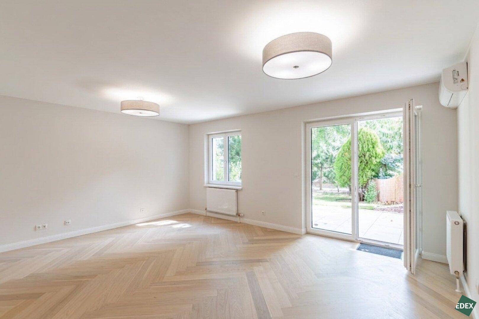 Geräumiges, unmöbliertes Wohnzimmer mit hellem Holzboden und großem Fenster zum Garten.