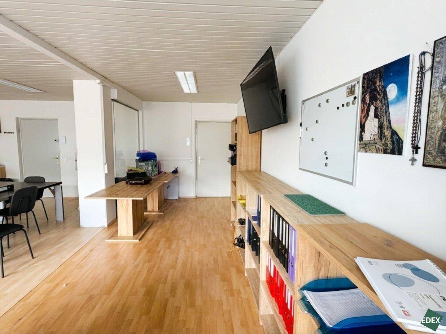 Ansicht eines offenen Bürobereichs mit Holzregalen, einem Wandfernseher und einem großen Holztisch als Arbeitsfläche.