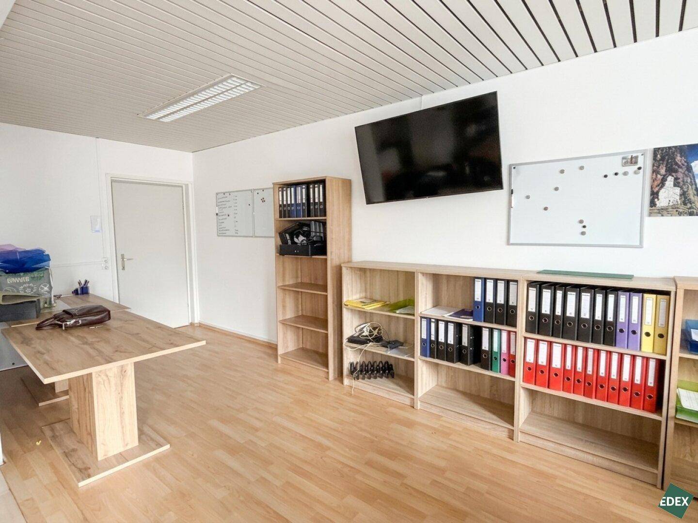 Weitere Perspektive des Büros mit Holzregalen, einem großen Holztisch und einem an der Wand befestigten Bildschirm.