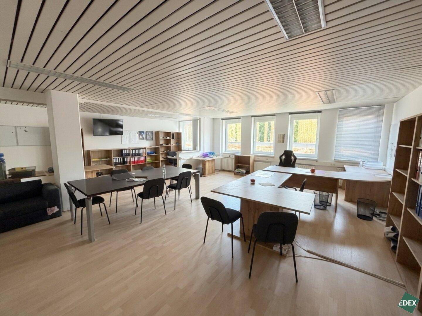 Weitläufiges Büro mit verschiedenen Arbeitsbereichen, einem Fernseher und viel Tageslicht durch große Fenster.