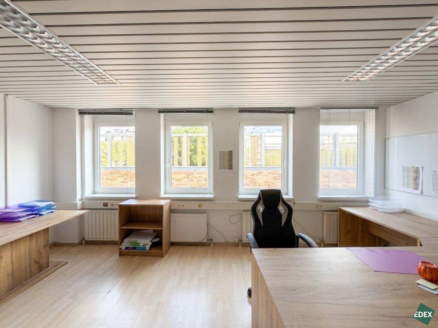 Detailansicht eines Bürobereichs mit großen Fenstern, Heizkörpern und hellem Laminatboden, der viel Licht hereinlässt.