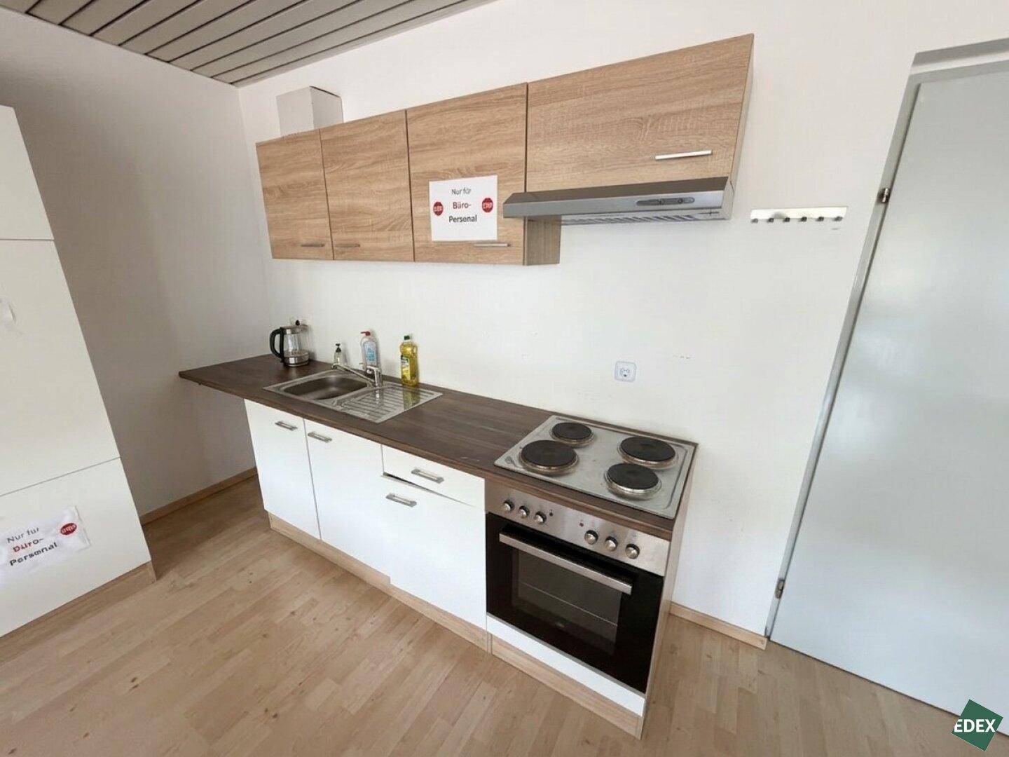 Funktionale Küchenzeile mit Herd, Backofen, Spüle und Holzschränken in einem Bürobereich, praktisch und sauber.