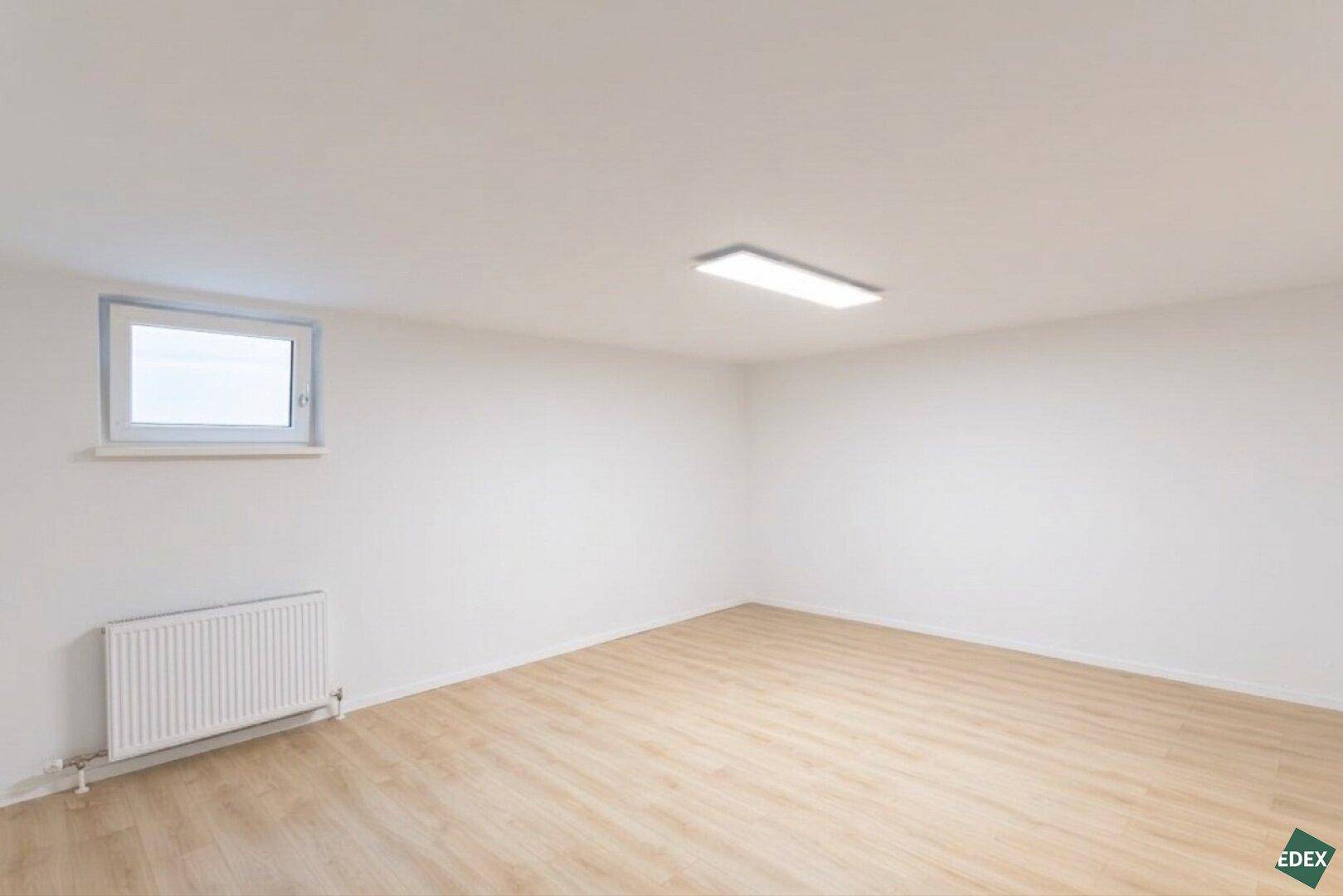 Leerer Raum mit hellem Laminatboden, weiß gestrichenen Wänden und einem Fenster.
