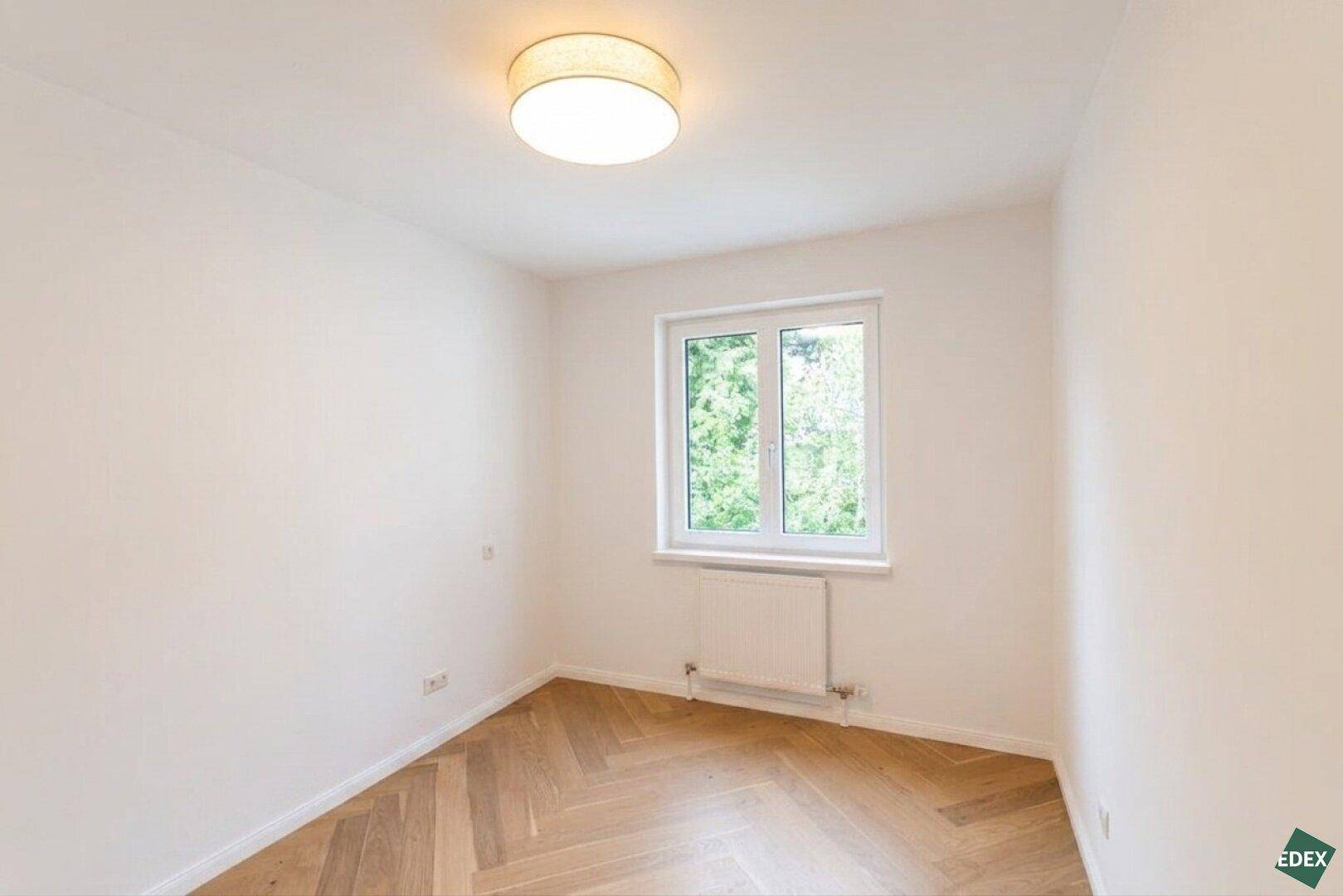 Helles, unmöbliertes Zimmer mit Holzboden und Fenster mit Blick ins Grüne.