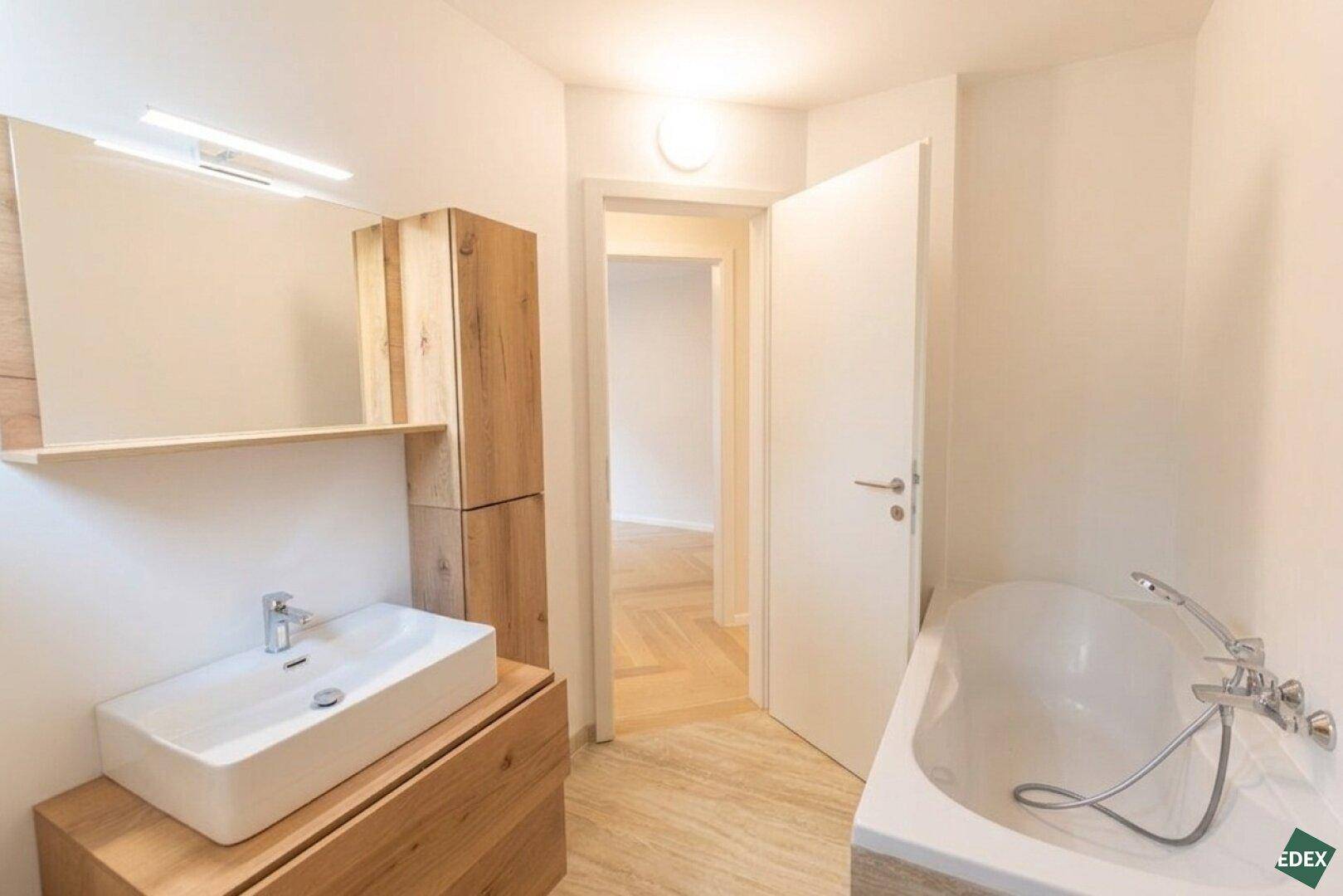 Modernes Badezimmer mit Badewanne, Waschbecken und Holzmöbeln, in hellen Farben.