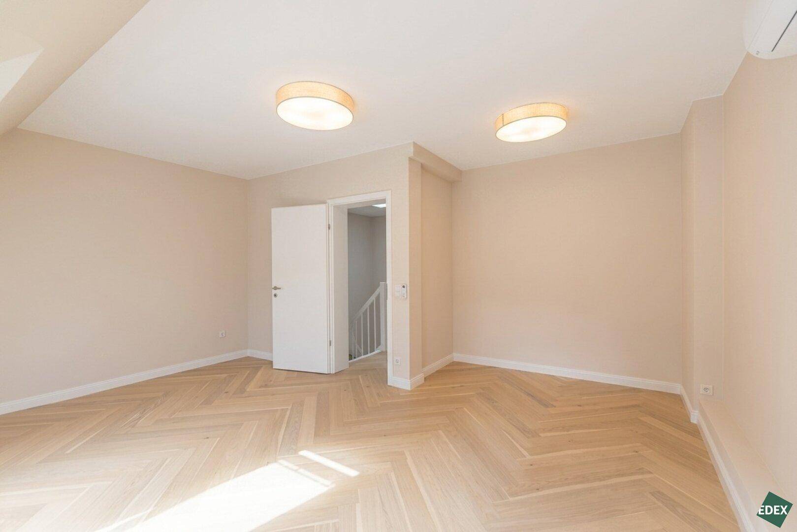 Geräumiges, unmöbliertes Zimmer mit Holzboden und zwei Deckenleuchten.